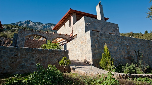 Un bâtiment en pierre avec un toit rouge et une cheminée, entouré de verdure et adossé à un paysage montagneux.