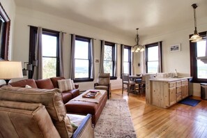 A spacious living room with a brown leather sofa, a dining area with a table and chairs, and a kitchen with wooden cabinets and a large window.