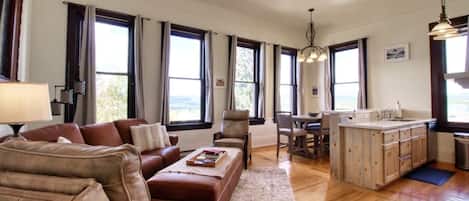 A spacious living room with a brown leather sofa, a dining area with a table and chairs, and a kitchen with wooden cabinets and a large window.