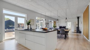 Eine moderne Küche mit einer großen Kochinsel, einem Essbereich mit einem Holztisch und Stühlen sowie Blick ins Freie durch große Fenster.