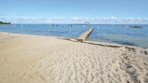Ein sandiger Strand mit einer hölzernen Pier, die sich in ruhiges, blaues Wasser erstreckt.