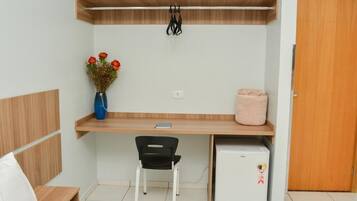 Um quarto pequeno com uma cama, uma escrivaninha com cadeira, um frigorífico pequeno, um vaso com flores e uma porta de madeira.