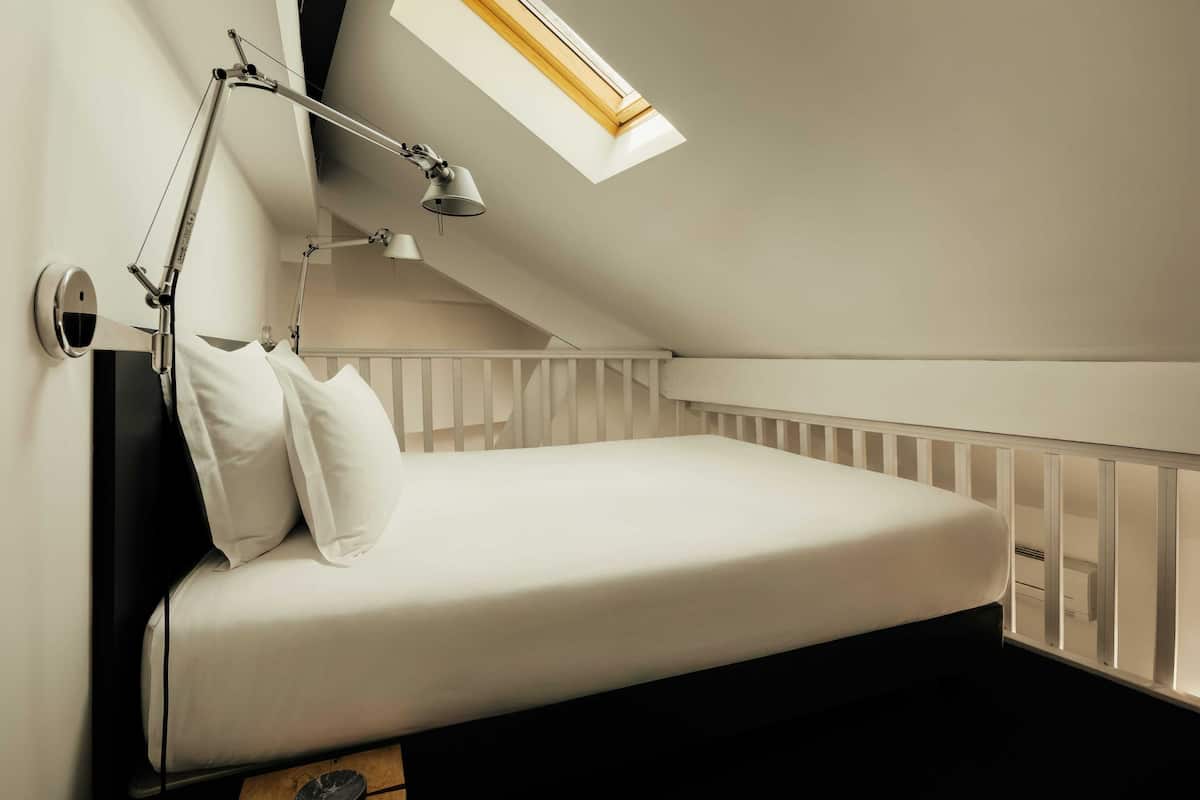 Une petite chambre avec un lit simple, un plafond en pente, un puits de lumière, une lampe de bureau et les barreaux blancs d’un berceau.