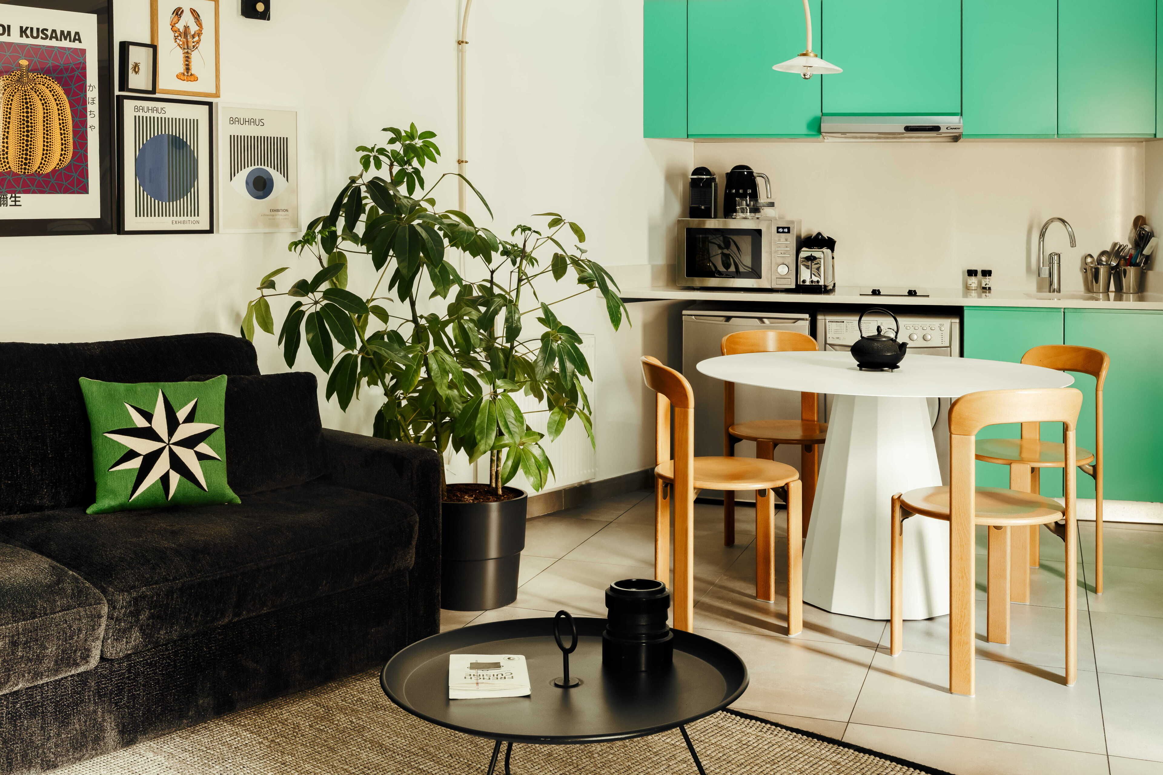 Una cocina moderna con armarios azul verdoso, una mesa de comedor y sillas. Hay un sofá negro con un cojín verde y una planta en maceta.