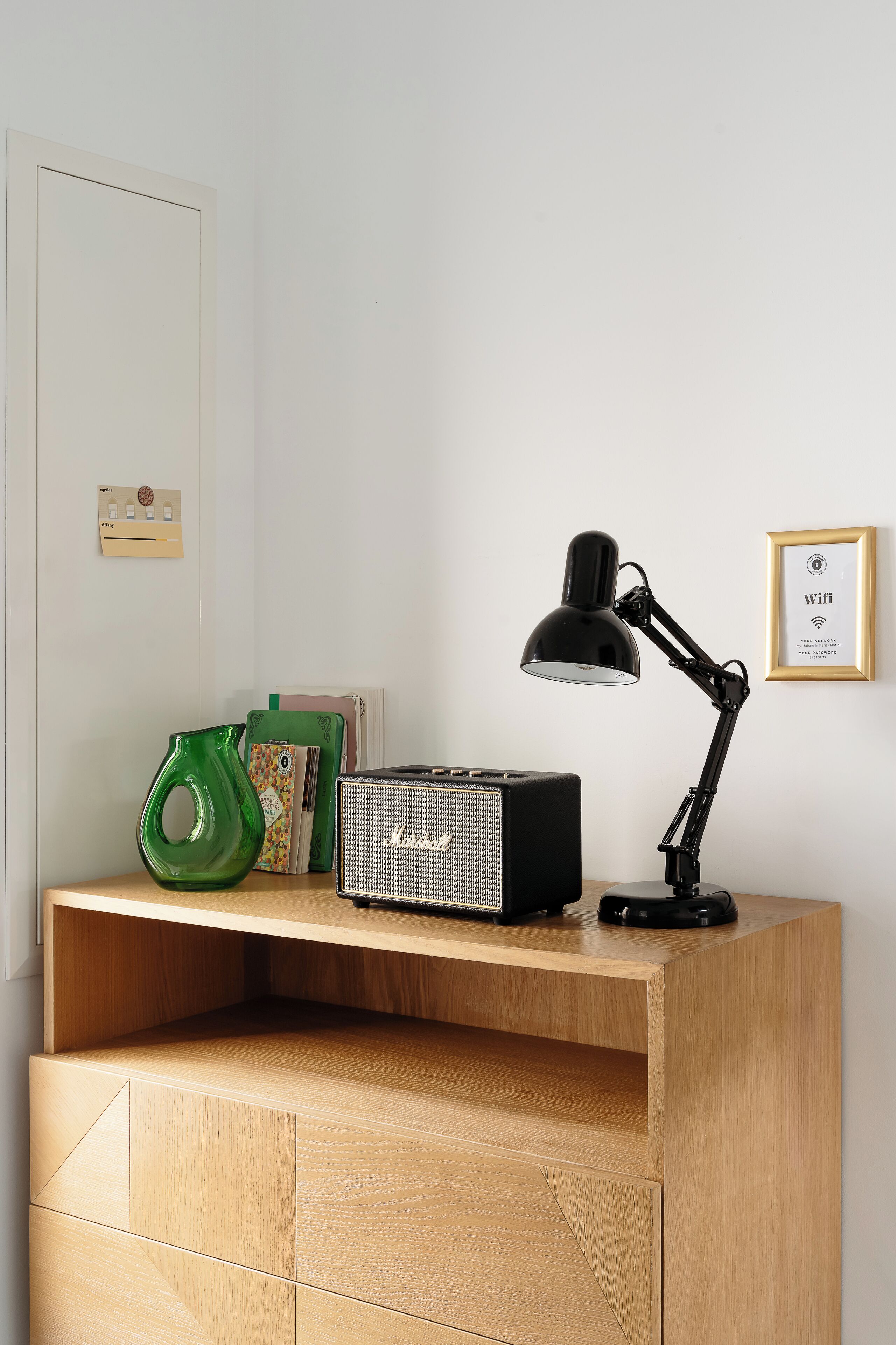 Un bureau en bois, sur lequel se trouvent une lampe de bureau noire, un vase vert et un tableau encadré accroché au mur.