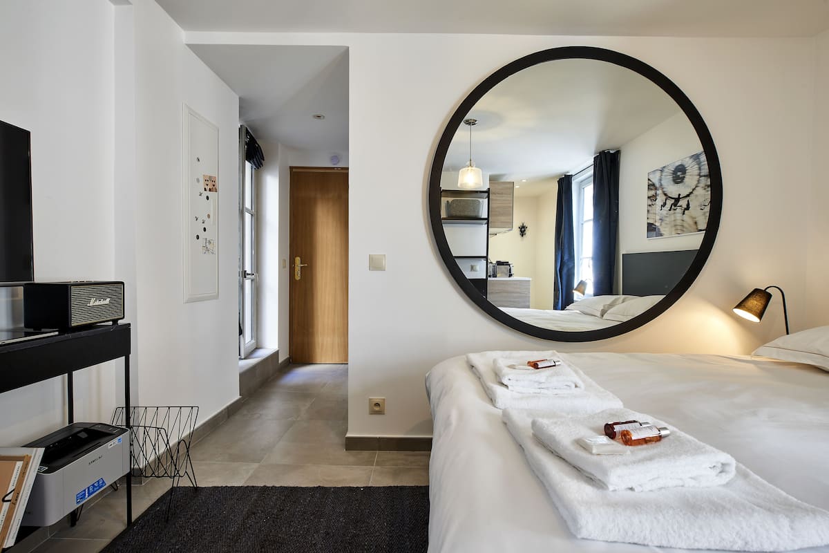 Une chambre d’hôtel avec un grand miroir rond, un lit avec du linge de lit blanc, un bureau avec une imprimante et une vue sur le couloir.