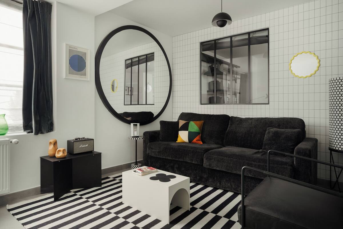 Un salon moderne avec un tapis rayé noir et blanc, un canapé noir agrémenté de coussins colorés, une table basse blanche, une petite table d’appoint noire avec un haut-parleur, et un grand miroir accroché au mur.
