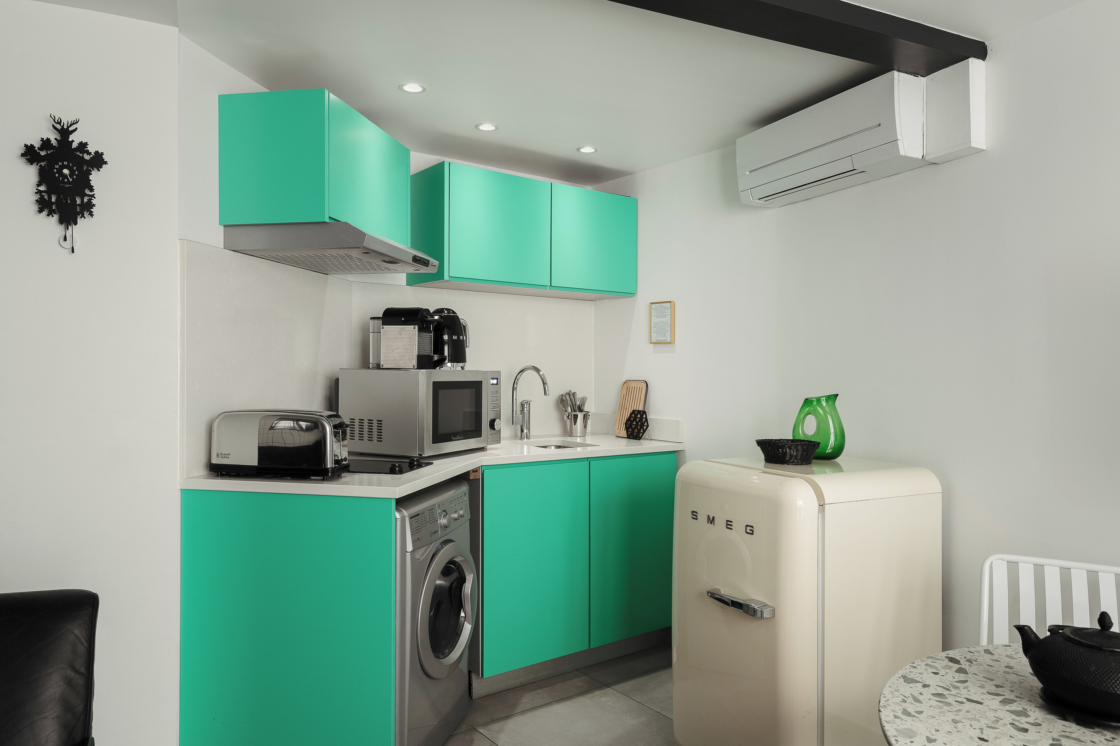 A compact kitchen with teal cabinets, a washing machine, a microwave, a toaster, and a vintage-style refrigerator.