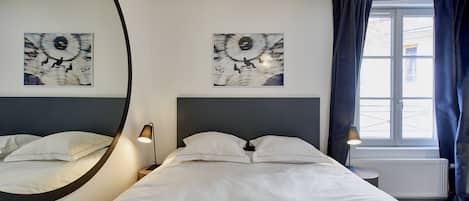 A hotel room with a large bed, two wall-mounted artworks, a round mirror, a bedside table with a lamp, and a window with curtains.