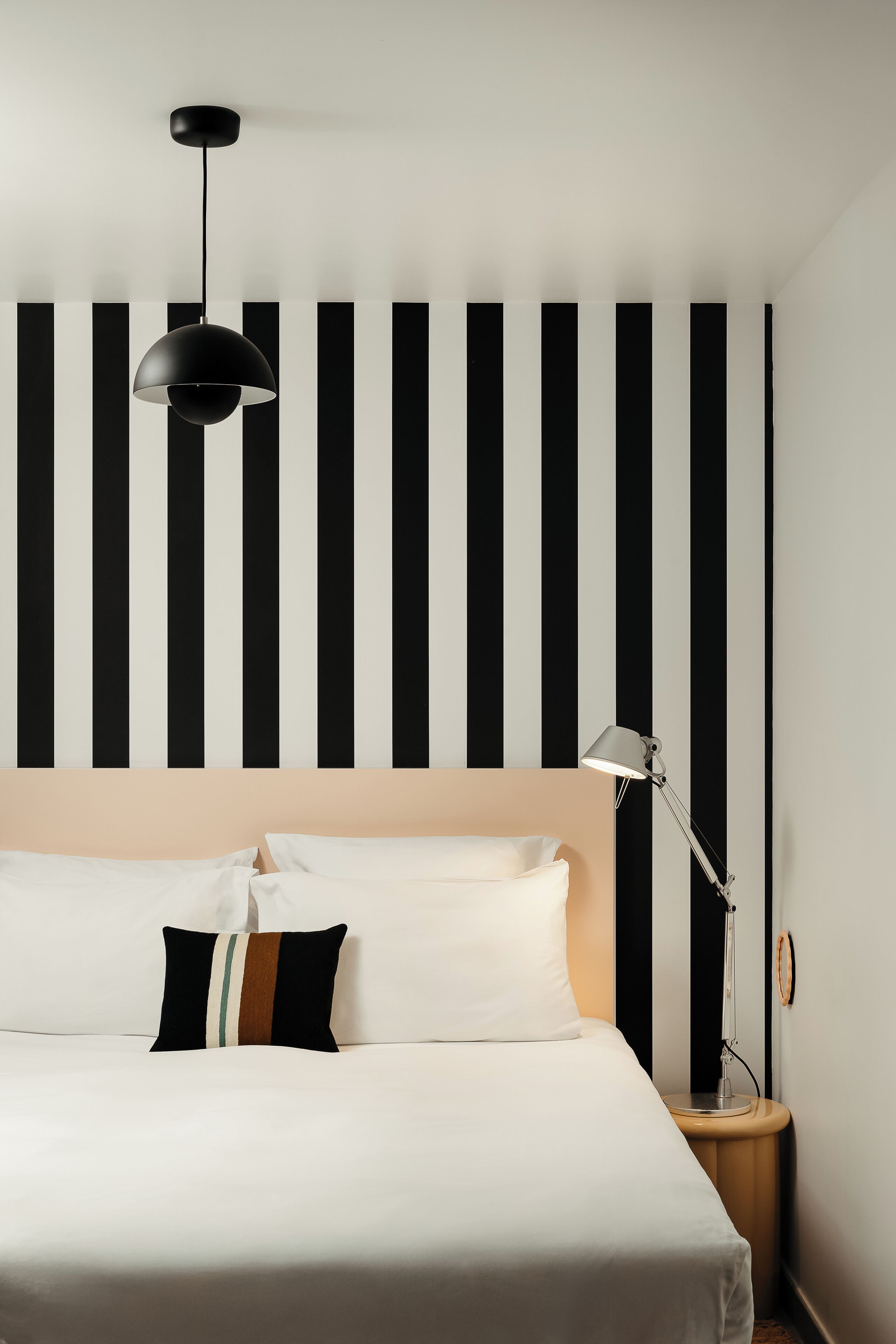 Un lit bien fait, avec du linge de lit blanc et un seul oreiller, une lampe de chevet et un mur orné de rayures verticales noires et blanches.