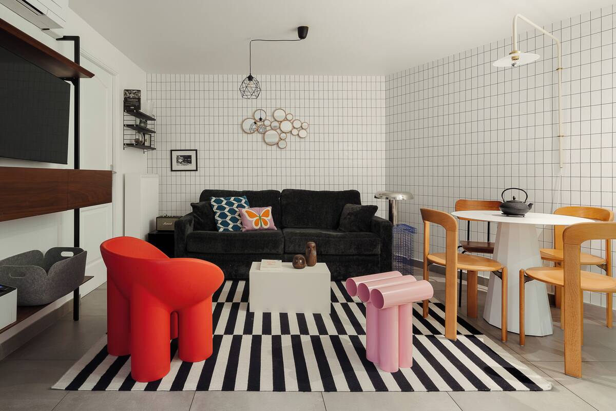Un salon moderne avec un canapé noir, des tabourets rouges et roses, une table basse blanche et un coin repas comprenant une table et des chaises en bois.