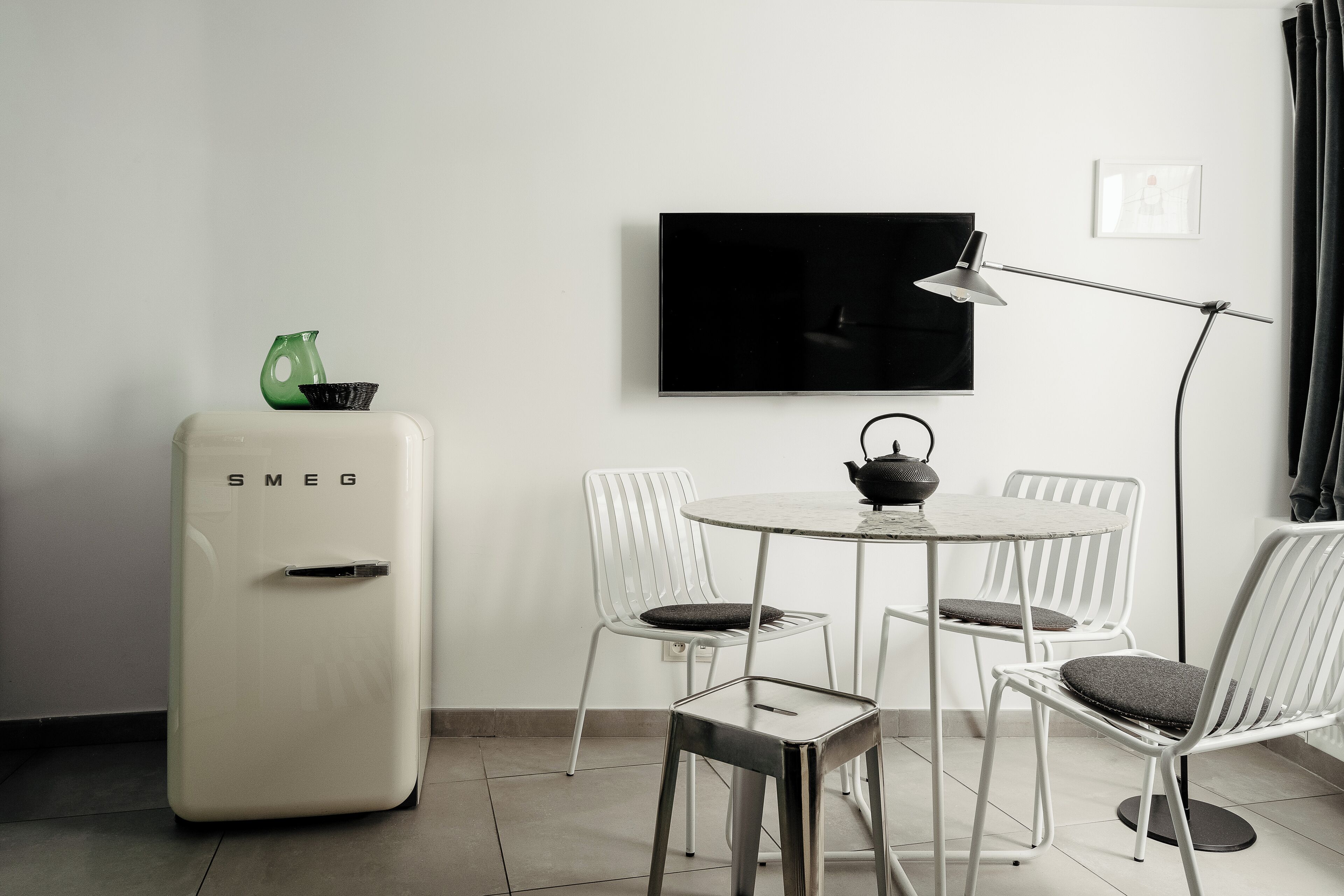Habitació moderna amb nevera blanca, televisió de pantalla plana penjada a la paret, taula rodona de menjador amb cadires blanques i una làmpada de peu.