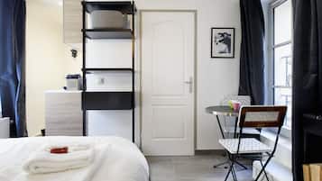 A hotel room with a bed, a desk with a chair, a radiator, a door, and a window with curtains.