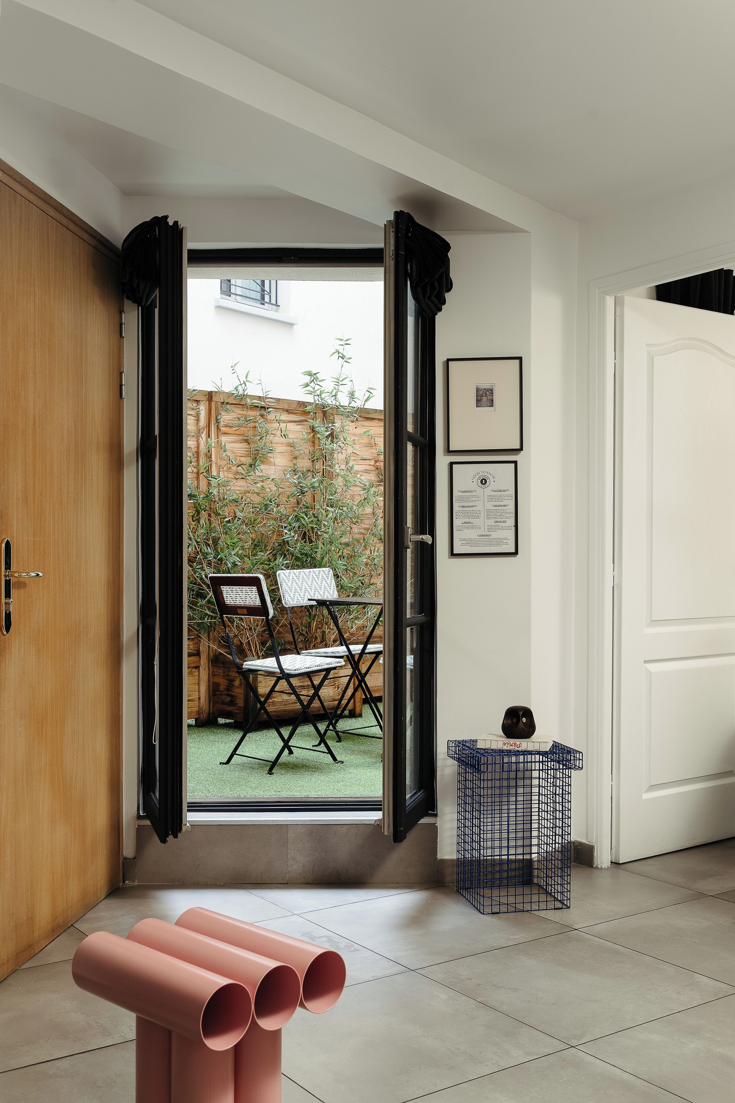 Una habitación con puerta de madera, una mesa pequeña con objetos cilíndricos rosas, una cesta de mimbre azul y vistas a un patio exterior con sillas y una valla de bambú.