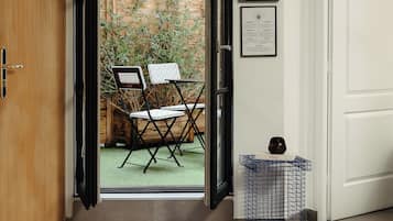 A room with a wooden door, a small table with pink cylindrical objects, a blue wire basket, and a view of an outdoor patio with chairs and a bamboo fence.