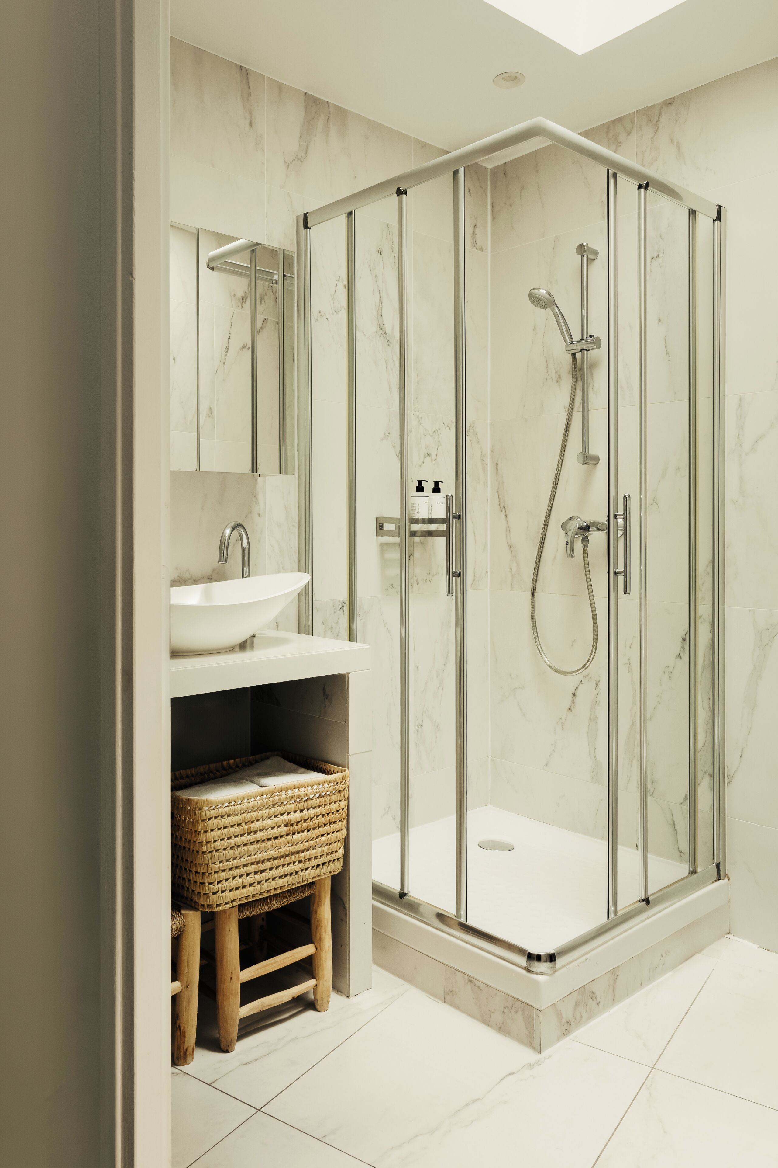 Een moderne badkamer met een douchecabine van glas, een witte wastafel en een rieten kruk.