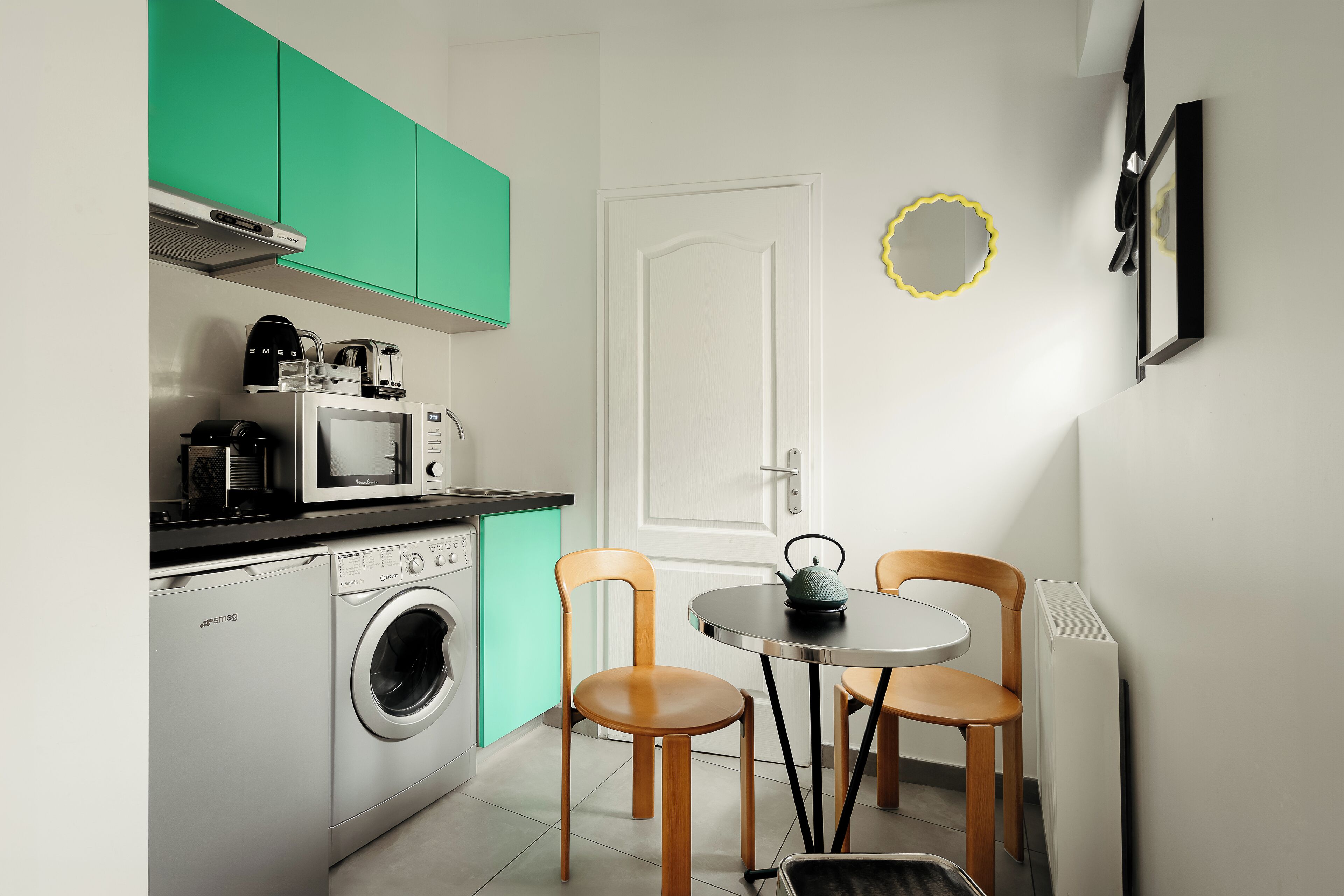 Cocina compacta con lavadora, microondas y cafetera. Se aprecia una mesa de comedor con sillas y una alfombra pequeña. Se ve una puerta blanca y un adorno amarillo.