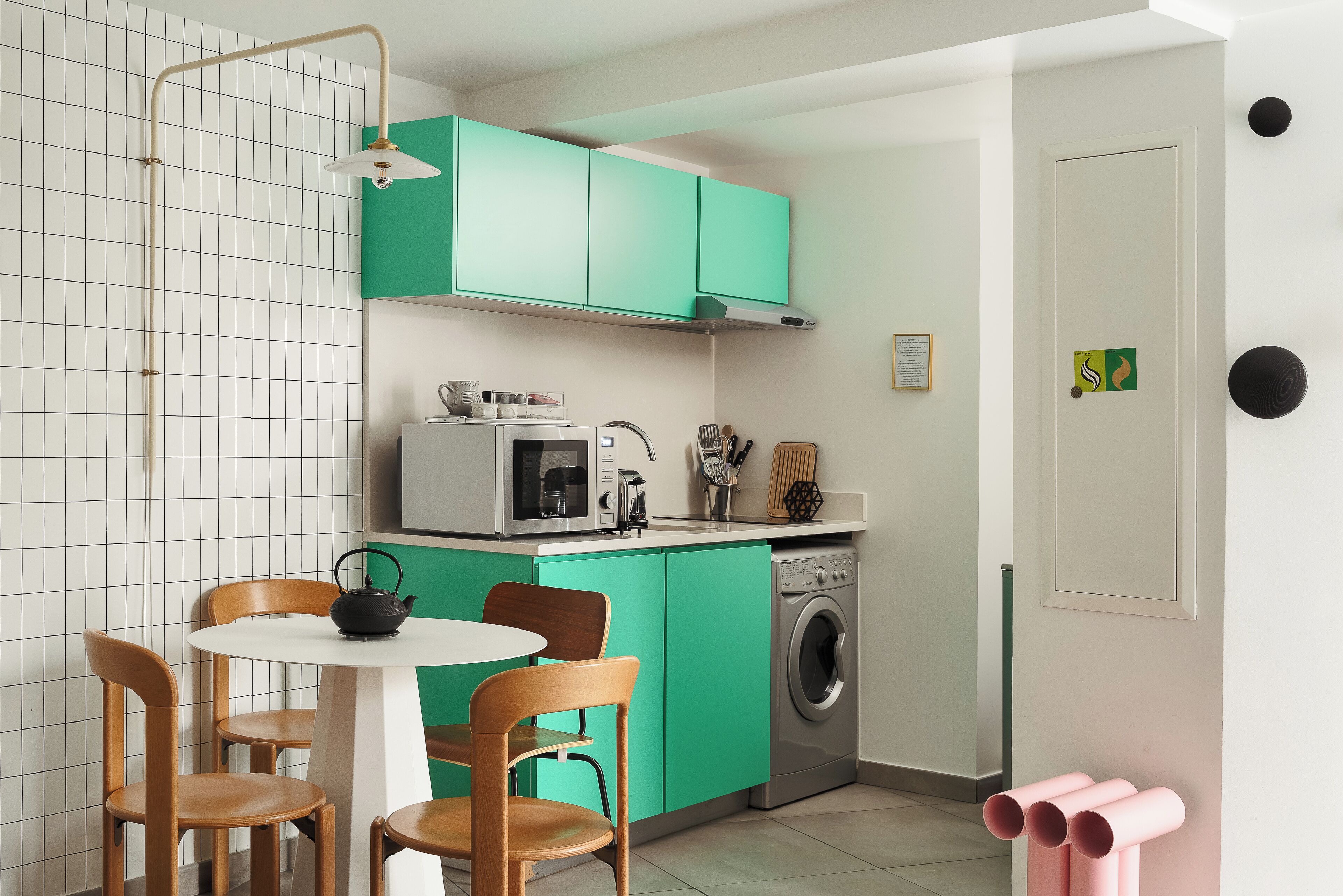 Una cucina compatta con mobili color petrolio, un forno a microonde, una macchina da caffè e una lavatrice. Si nota un tavolo rotondo con sedie e un piccolo bollitore nero.