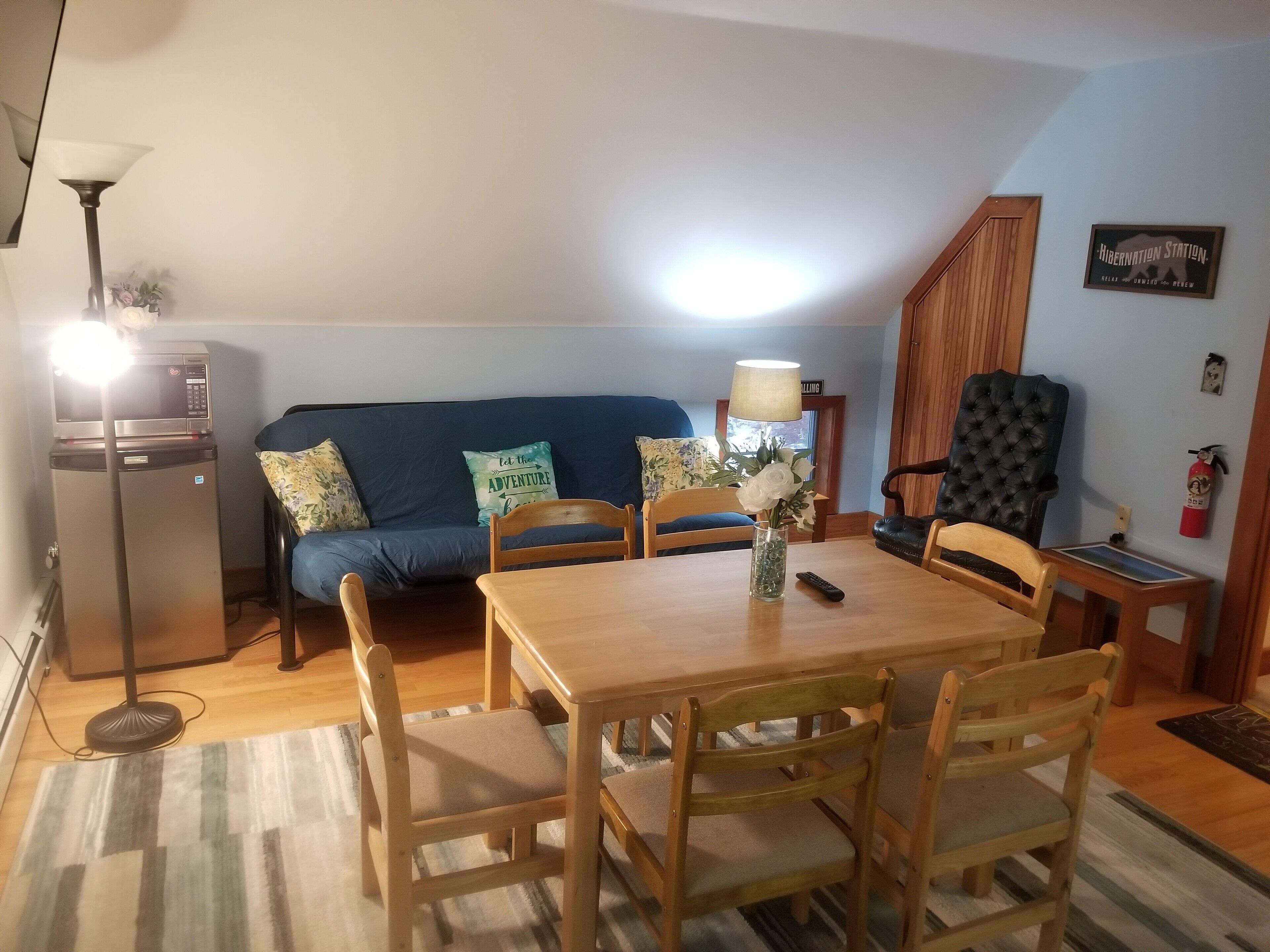 Un salon chaleureux avec un canapé bleu, une table à manger en bois et des chaises. On aperçoit un micro-ondes sur la gauche, une lampe sur la droite et un vase rempli de fleurs sur la table.