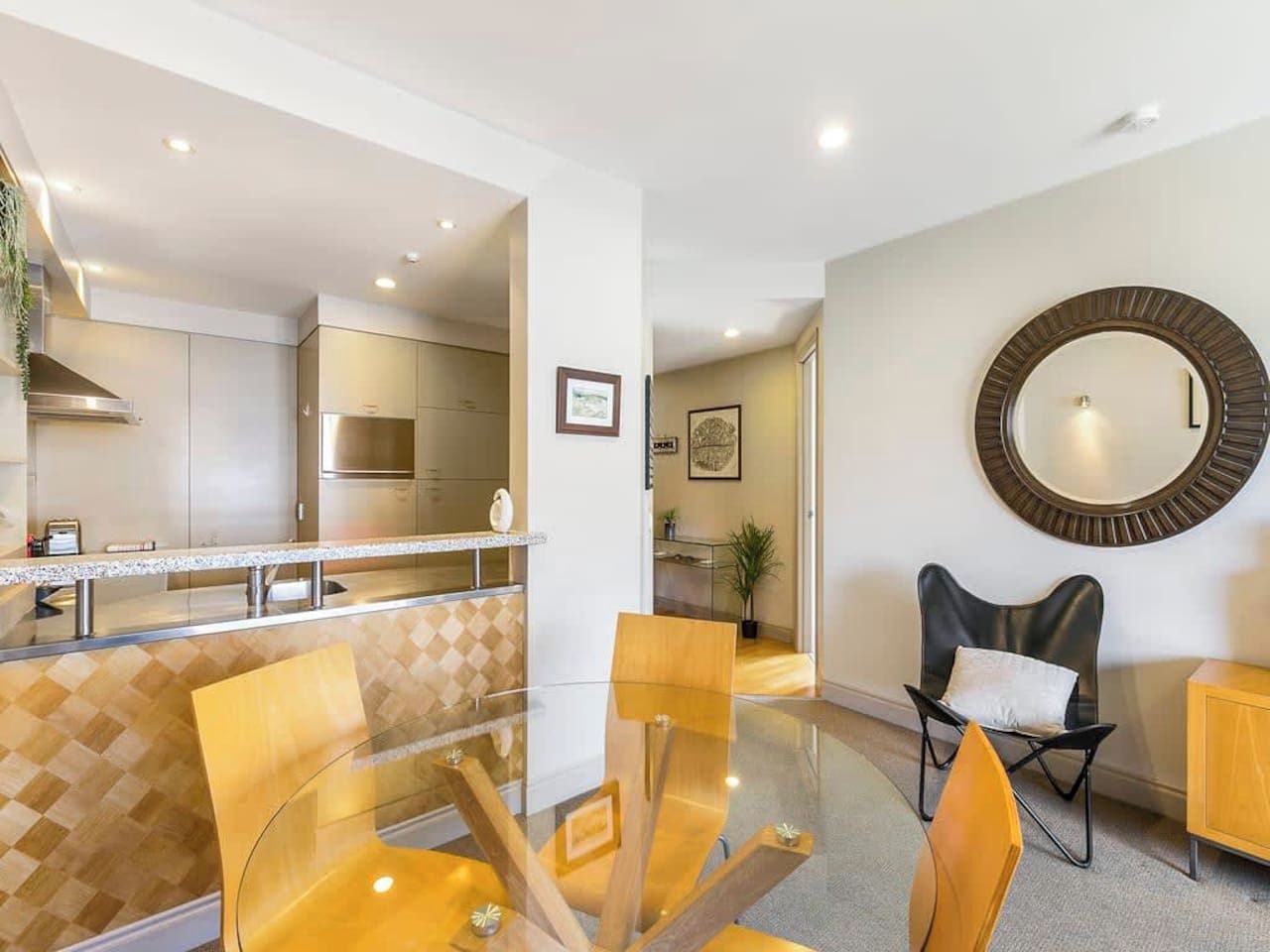 Une cuisine moderne avec une table et des chaises en verre, un miroir rond accroché au mur et une vue sur le salon attenant.