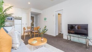 A modern living room with a sofa, a glass coffee table, a TV on a glass stand, a dining area with chairs, and a kitchen with white cabinets and appliances. - Cozy City Apt with Pool Gym & View by Zodiak Stays (Auckland)