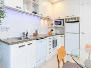 A modern kitchen with white cabinetry, a built-in microwave, a dishwasher, and a glass dining table with chairs. - Cozy City Apt | Pool, Gym & View | Zodiak Stays (Auckland)
