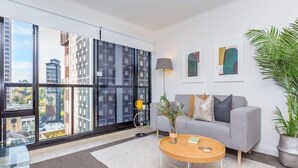 A modern living room with a grey sofa, a round coffee table, and a large window with a city view. - Cozy City Apt with Pool Gym & View by Zodiak Stays (Auckland)