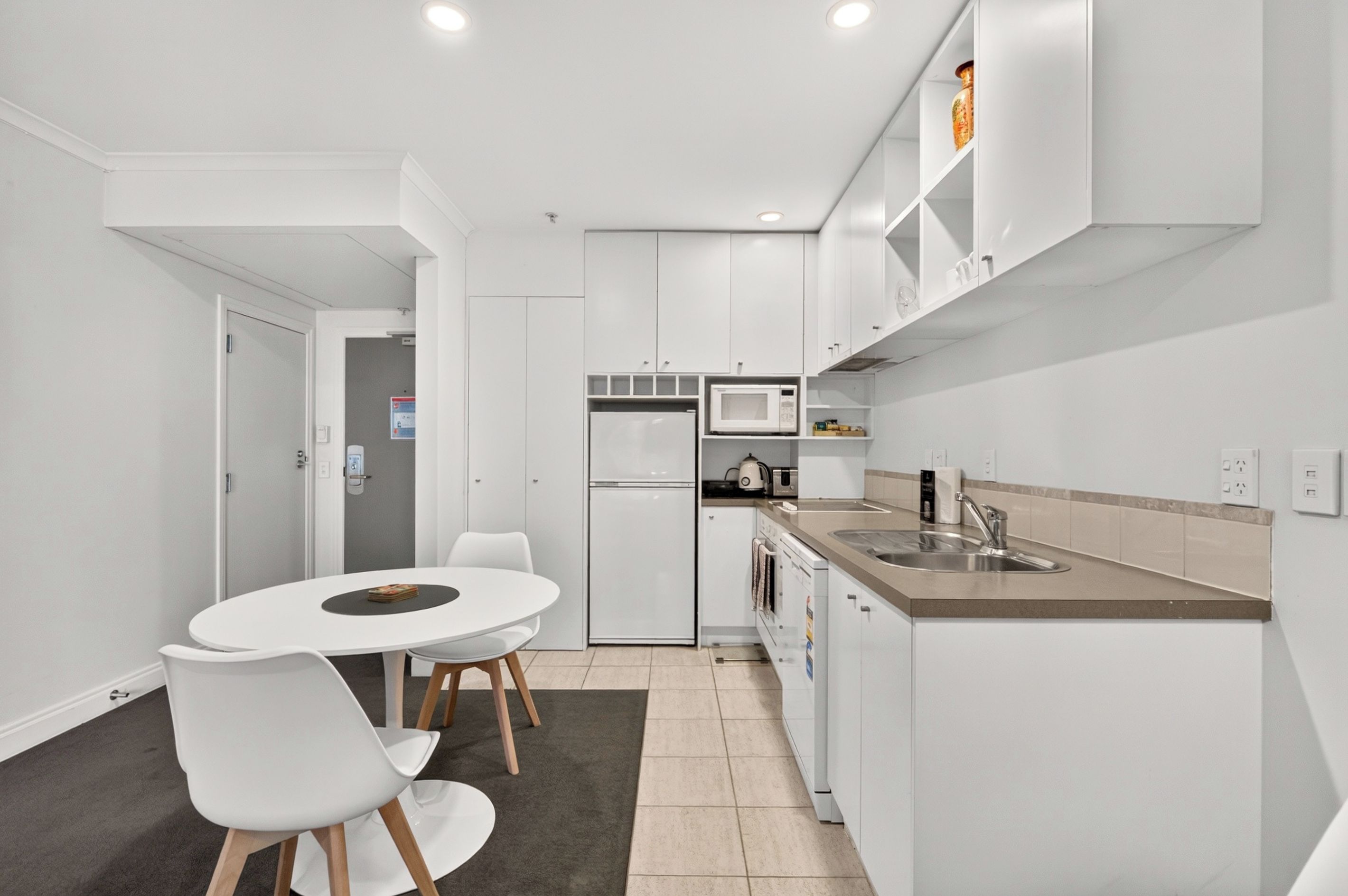 A compact kitchen with white cabinetry, a dining table with chairs, a microwave, a refrigerator, and a sink.