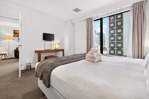 A hotel room with a large bed, a desk with a flat-screen TV, a chair, a small trash bin, and a view of the city through the window. - Superking Bed in Stylish Apartment I Zodiak Stays (Auckland)