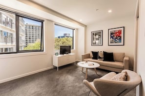 A modern living room with a sofa, a coffee table, and a TV on a white cabinet. There are two windows with a view of buildings and trees. - Park Views in CBD Next to Myers Park by Zodiak (Auckland)