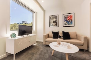 A modern living room with a television on a white cabinet, a beige sofa with black cushions, a white coffee table, and two framed pictures on the wall.