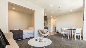 A modern hotel room with a bed, a round dining table with chairs, a sofa, and a small white table with a decorative item. - Beautiful 2BR Unit next to Myers Park Zodiak Stays (Auckland)