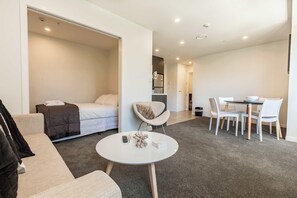A modern hotel room with a bed, a round dining table with chairs, a sofa, and a small white table with a decorative item.