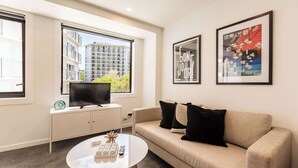 A modern hotel room with a sofa, a coffee table, a TV on a cabinet, and large windows offering a view of a building outside. - Beautiful 2BR Unit next to Myers Park Zodiak Stays (Auckland)