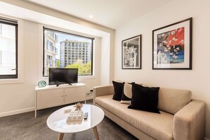 A modern hotel room with a sofa, a coffee table, a TV on a cabinet, and large windows offering a view of a building outside.