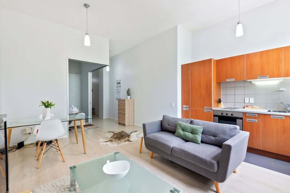 A modern living room with a glass dining table, a grey sofa, and a kitchen with wooden cabinets.