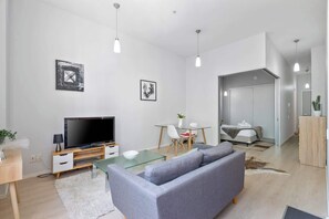 A modern living room with a television, sofa, dining table, and chairs. There is a separate bedroom with a bed and a wardrobe.