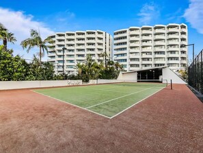 Un court de tennis à surface rouge, entouré d’une clôture blanche et situé à côté d’un immeuble à plusieurs étages doté de balcons.