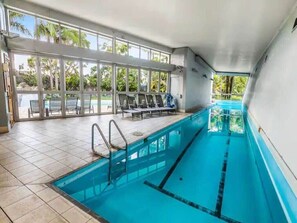 An indoor swimming pool with clear glass walls, a tiled floor, and a poolside area with chairs. - Lovely Apartment - Gym & Parking by Zodiak Stays (Auckland)