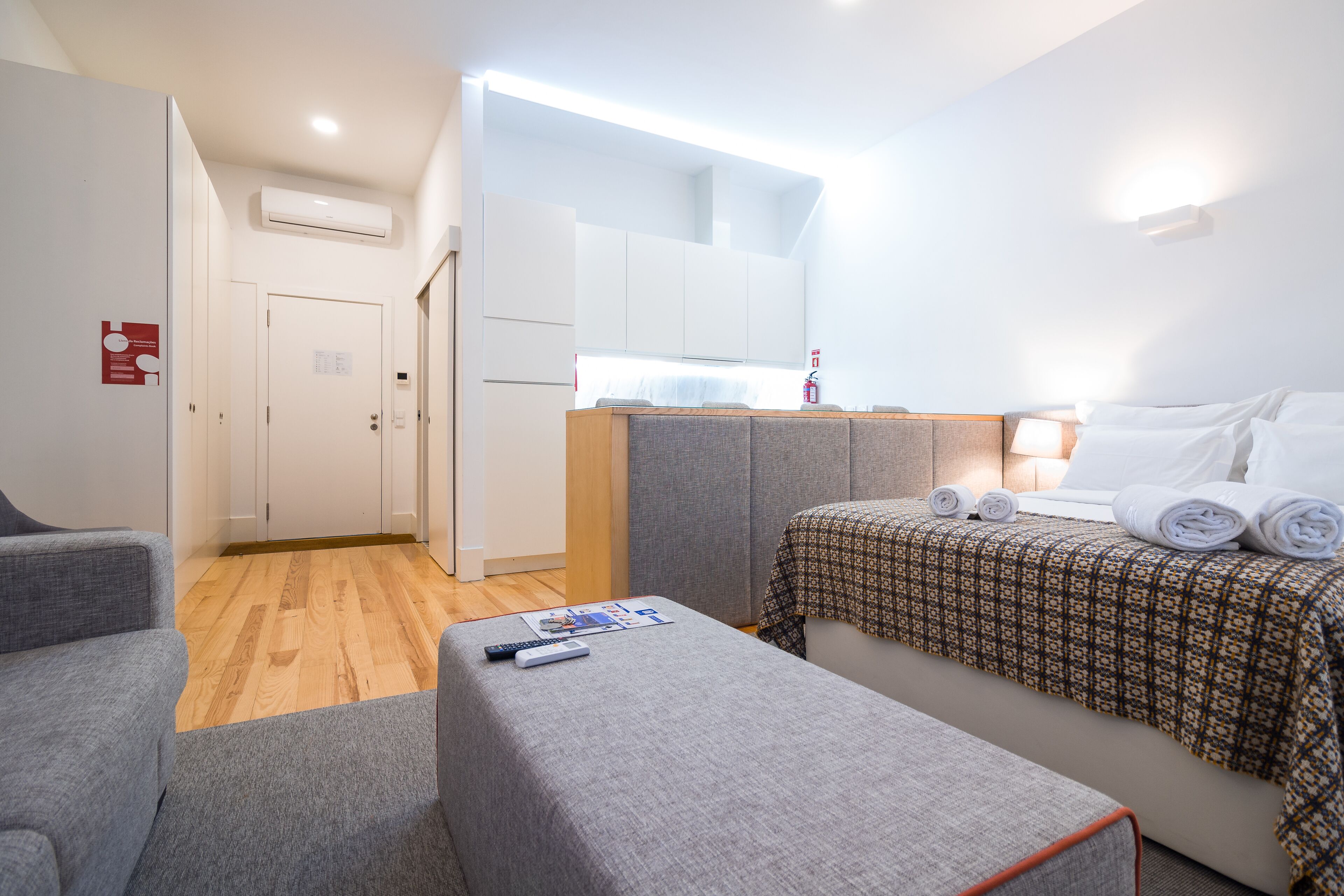 A compact hotel room with a kitchenette, a bed with a patterned bedspread, a grey sofa, and a small table with a remote control.