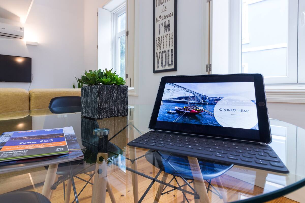 Una habitación moderna con una mesa de vidrio, una computadora portátil mostrando una vista panorámica, un sofá, una planta y una cita enmarcada en la pared.