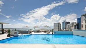 En pool på taket med solstolar och parasoller, omgiven av höga byggnader.
