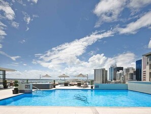 Une piscine sur le toit, agrĂ©mentĂ©e de chaises longues et de parasols, et entourĂ©e dâimmeubles de grande hauteur.