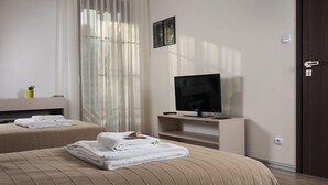 A hotel room with two beds, a TV, a small shelf, and a window with sheer curtains.