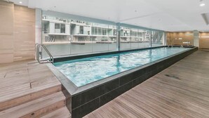 An indoor swimming pool with a wooden deck, glass walls, and a modern design. - Beautiful 2BR - Pool, Gym & Parking Zodiak Stays (Auckland)