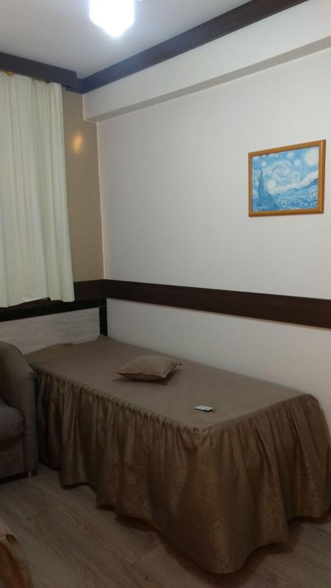 A single bed with a brown bedspread, a wooden headboard, a white wall with a framed picture, and a window with white curtains.