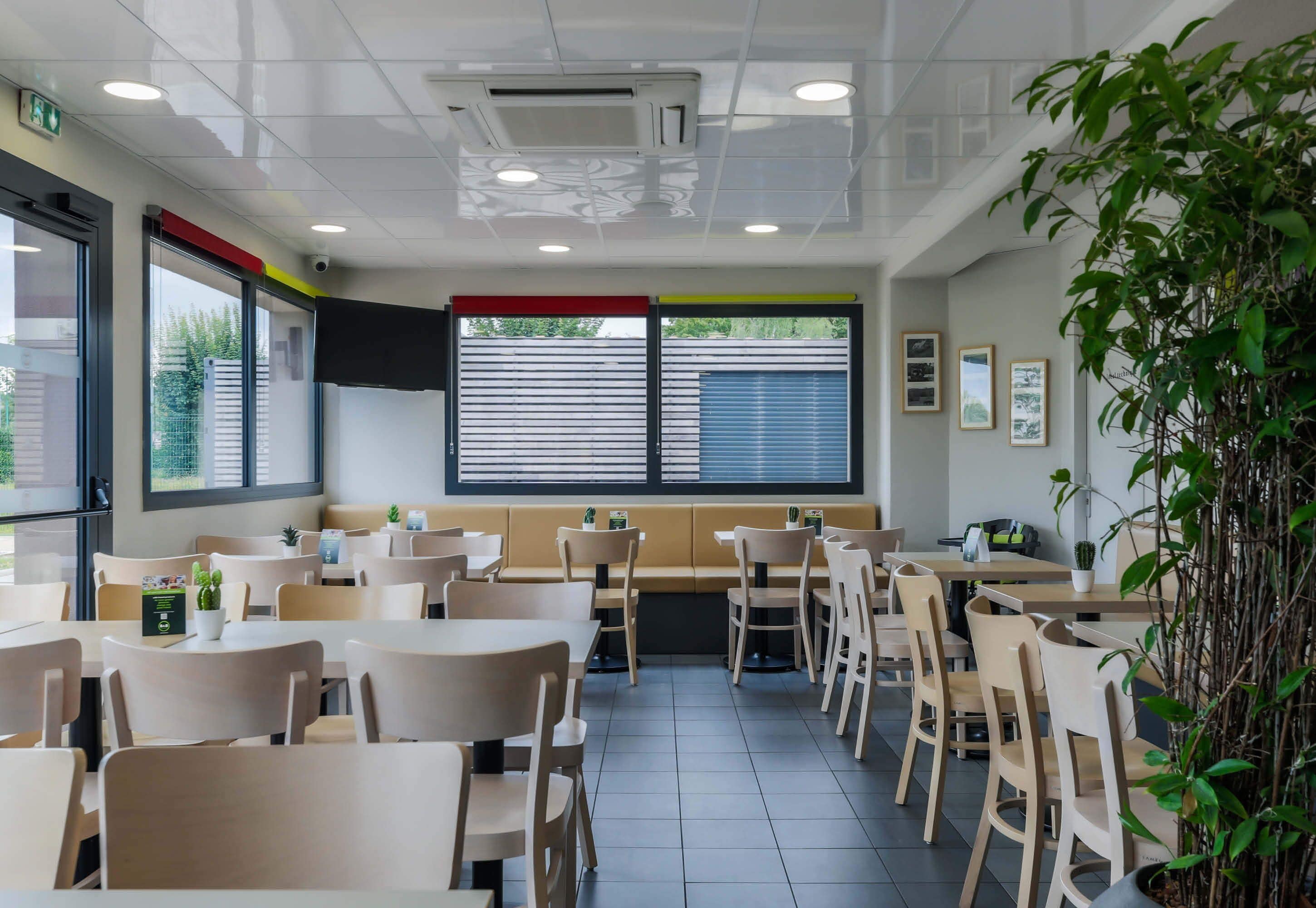 A restaurant interior with tables and chairs, a large window with blinds, and a plant on the right.