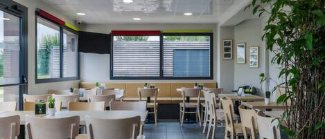 Interior de restaurante con mesas y sillas, un ventanal con persianas y una planta a la derecha.