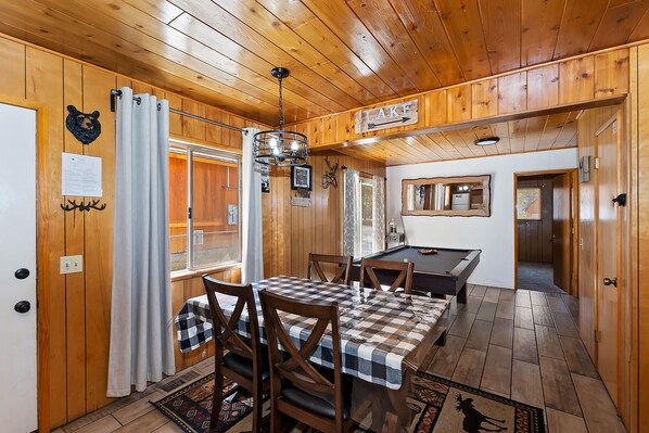 Interior de una cabaña de madera con una mesa de comedor, sillas, una mesa de billar y una zona de cocina.