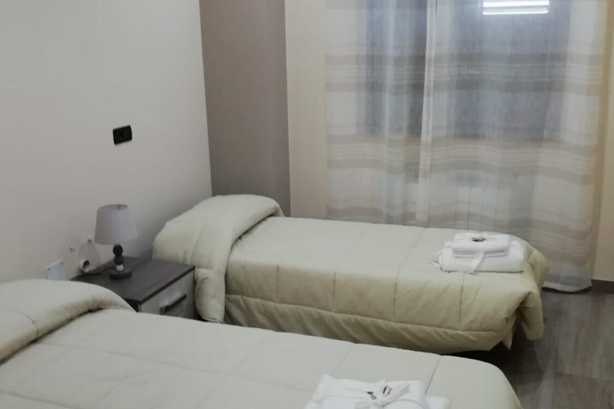 A hotel room with two beds, a nightstand with a lamp, and a window with a curtain.